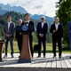 G7 leaders are shown standing on a stage with mountains in the background.